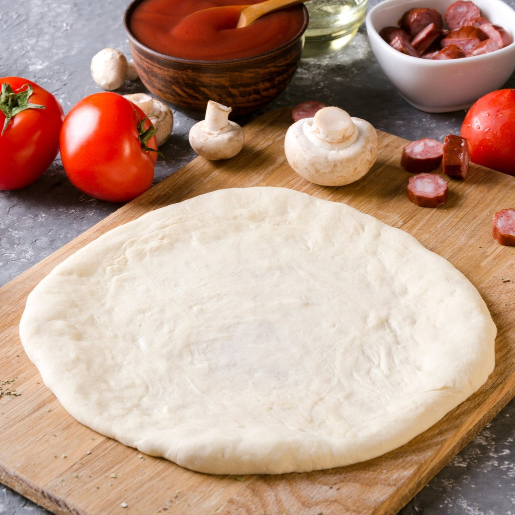 Pizza dough on a wooden cutting board with vegetables around it